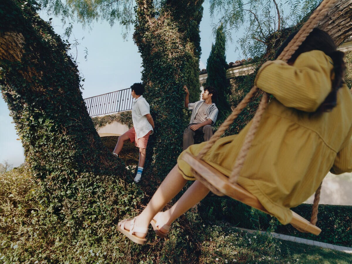 Three children are playing outside, facing away from the camera. One child is one a simple tree swing. 